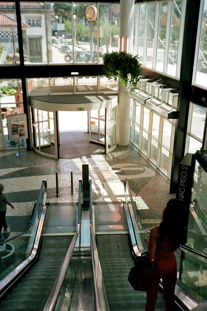 about-us-02 A woman descends an escalator towards a bright mall entrance with revolving doors and natural light.